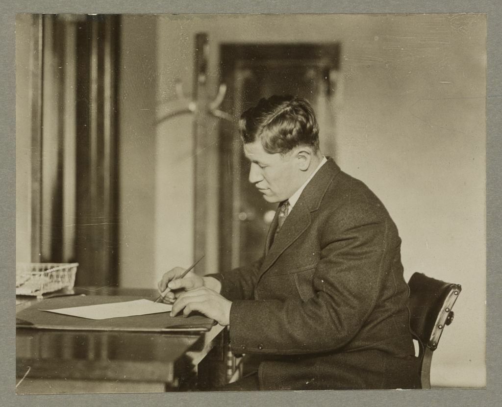 Photograph shows Jim Thorpe, half-length portrait, facing left, sitting at a desk, writing.