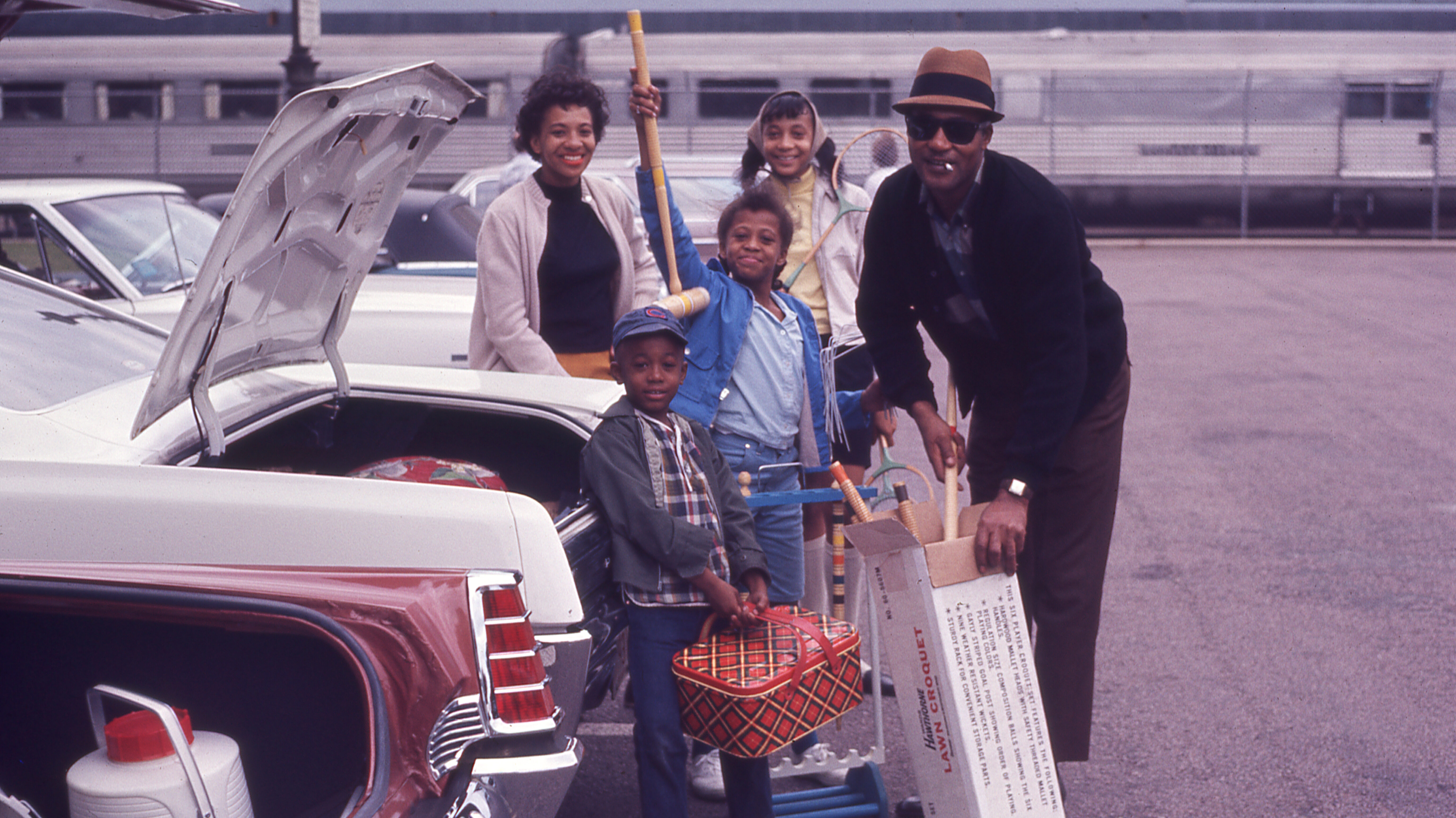 Family taking croquet game and picnic basket out of a trunk smiling, train in background.