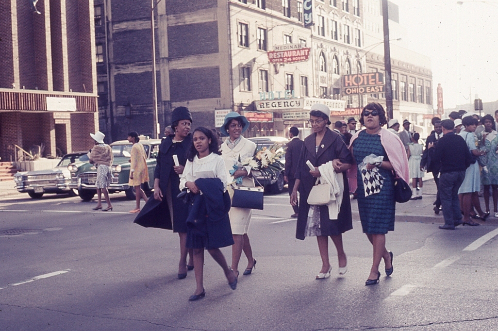Fashionable woman with child crossing city street, other pedestrians and traffic in background, sign.