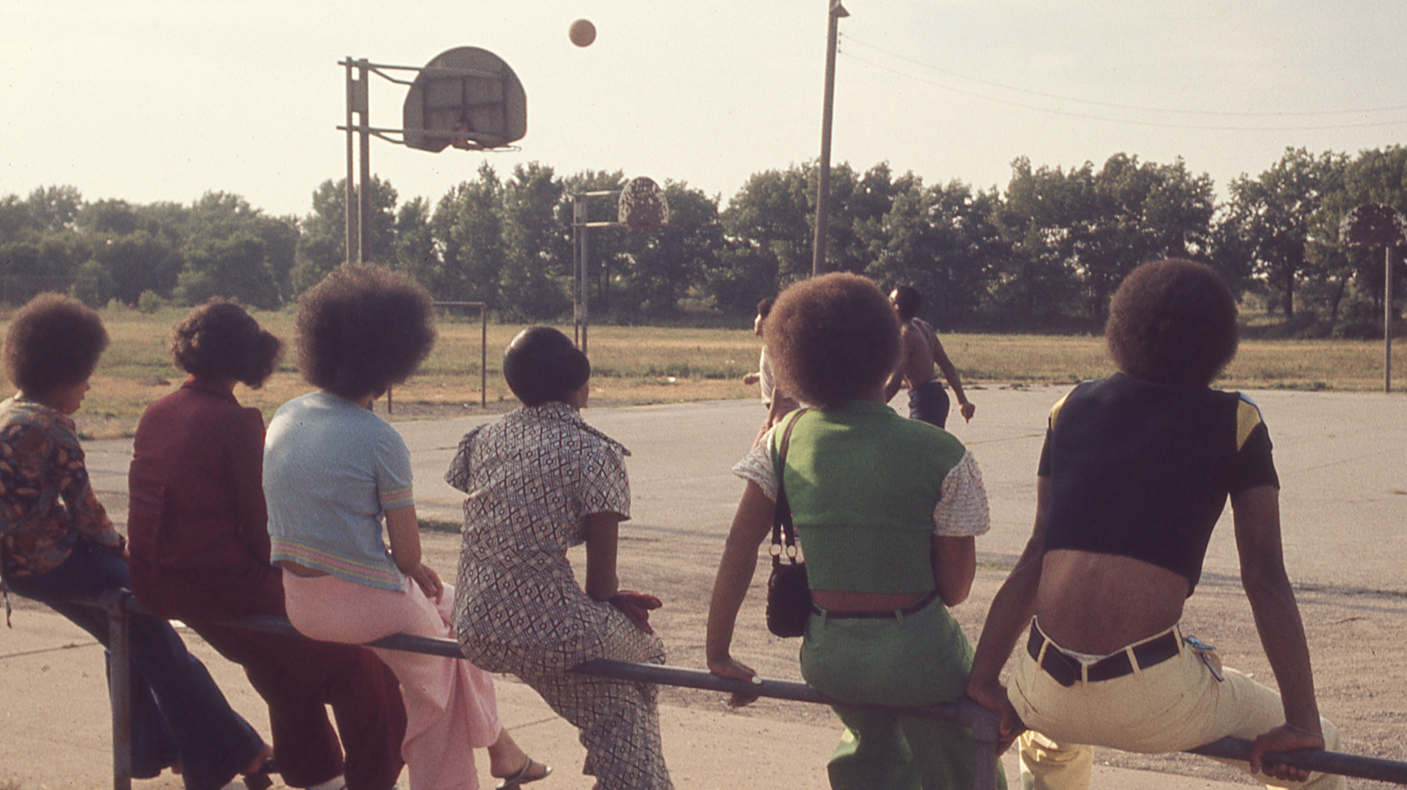 Teenagers/young adults with afros sitting together watching someone play basketball, viewed from behind.